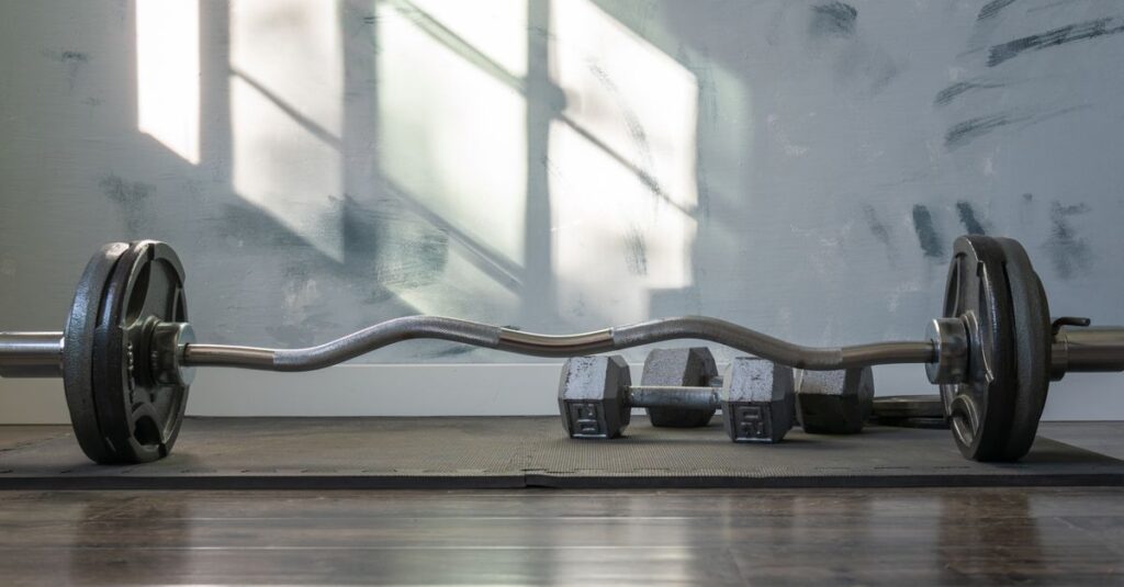 Barbell and dumbbells indoors on a gym mat with sunlight streaming through the window.
