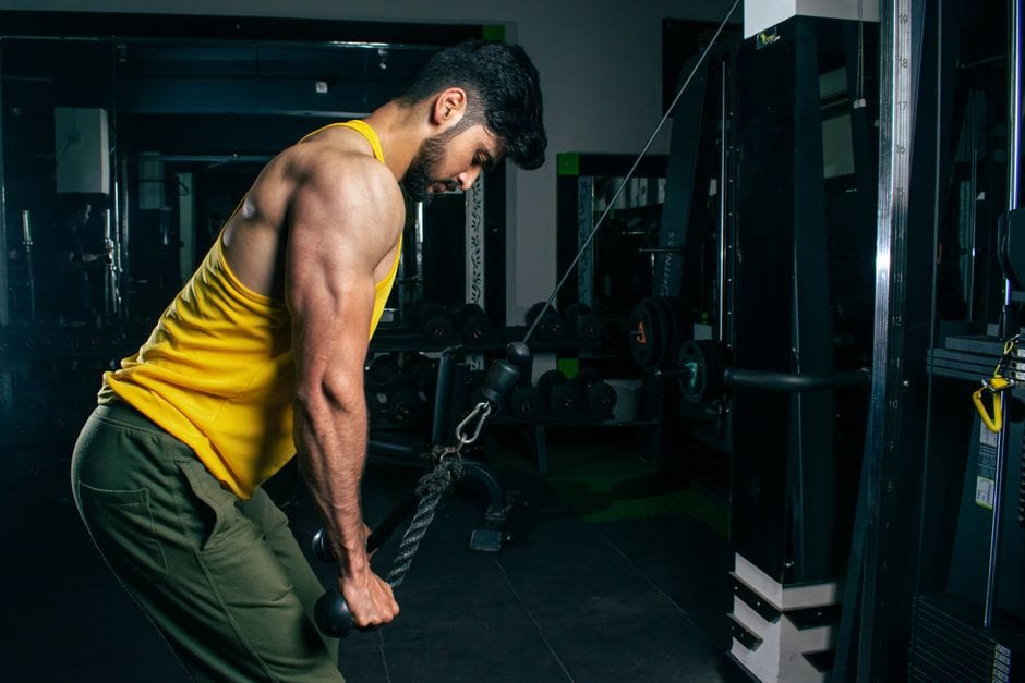 A fit man in a yellow tank top exercises using a cable machine in a gym setting.
